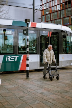 An elderly person using a walker crosses in front of a modern tram with the text 'RET' on its side. The tram is adorned with multiple colors including red, green, and white. In the background, there is a modern building with large glass windows.