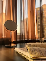 A round mirror stands on a polished table next to a notebook with handwritten notes. The scene is illuminated by warm sunlight streaming through sheer brown curtains. A glass with a small amount of liquid is placed nearby, and the urban skyline is faintly visible through the windows.