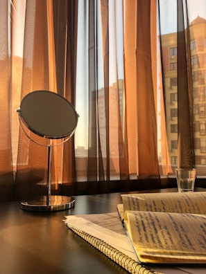 A round mirror stands on a polished table next to a notebook with handwritten notes. The scene is illuminated by warm sunlight streaming through sheer brown curtains. A glass with a small amount of liquid is placed nearby, and the urban skyline is faintly visible through the windows.