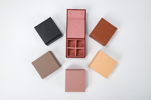 Several small, square boxes are arranged in a circle on a white background. These boxes are of various colors, including black, brown, beige, taupe, and pink. One box is open, revealing four compartments inside.