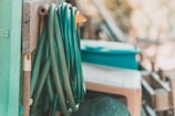 A garden hose is coiled neatly against a wooden panel next to a green container. Several other garden tools are partially visible, leaning against or hanging on the wood.