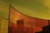 A series of tall, curved, tinted glass panels create an abstract and modern architectural feature. The panels have a strong reddish-orange tint, against a sky that is gradient from yellow to greenish. Shadows and reflections within the panels add depth and complexity to the scene.