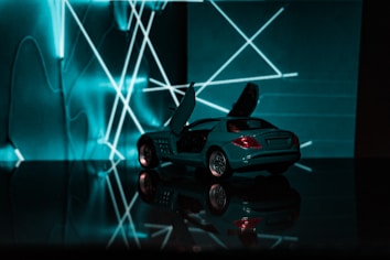 A small model car with open doors is placed on a glossy surface, reflecting its image. In the background, illuminated neon lights form geometric patterns, creating a dynamic and futuristic setting.