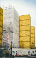 A construction site featuring stacked yellow shipping containers with scaffolding and a section covered in white tarp. In the foreground, there is a colorful mural with whimsical designs on the construction fence. Several street signs and traffic lights are also visible.