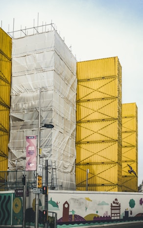 A construction site featuring stacked yellow shipping containers with scaffolding and a section covered in white tarp. In the foreground, there is a colorful mural with whimsical designs on the construction fence. Several street signs and traffic lights are also visible.