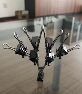 A sculpture made of black binder clips is arranged in a symmetrical and artistic formation resembling a tree or abstract shape, placed on a glass surface with reflections visible. The background includes indistinct furniture and a cardboard box, indicating an office or home setting.