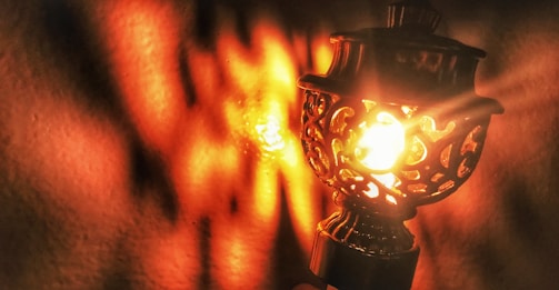An ornate metal lamp with intricate cut-out patterns emits a warm, glowing light. The light creates vibrant orange and yellow reflections and shadows on a nearby surface, enhancing the lamp's decorative details.