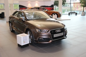 A sleek brown Audi A3 1.8 T quattro is prominently displayed inside a modern showroom. The car has a shiny surface and its stylish design is accentuated by the bright indoor lighting. There is a small informational stand in front of the vehicle, and another car is partially visible in the background, adding depth to the scene. Large windows in the showroom allow natural light to filter through, contributing to the polished and professional ambiance.