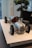A vintage silver race car is displayed on a white platform indoors. It features a classic design with an open cockpit, large steering wheel, and sleek body. The car has recognizable branding on the grille and is positioned next to a large potted plant.