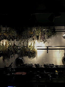 A dimly lit kitchen with bundles of dried herbs hanging on a rack above a tiled backsplash. Below, a counter holds various kitchen utensils, pots, and a cutting board. The lighting casts soft shadows on the tiles, creating a warm and rustic atmosphere.