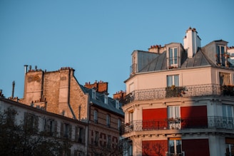 Several residential buildings with classic European architecture are depicted, showcasing stone facades, wrought iron balconies, and multiple chimneys. The structures are bathed in the warm glow of sunset or sunrise, casting soft shadows.
