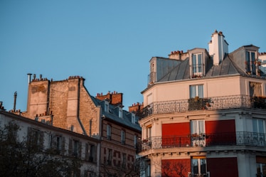 Several residential buildings with classic European architecture are depicted, showcasing stone facades, wrought iron balconies, and multiple chimneys. The structures are bathed in the warm glow of sunset or sunrise, casting soft shadows.