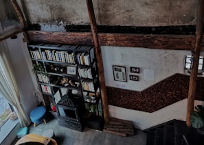 A cozy interior space with rustic and vintage design elements, featuring a black metal bookshelf filled with books and decorated with plants. The walls exhibit a weathered texture, and there are wooden beams adding to its rustic charm. A small blue stool and a fireplace with decorative items are placed nearby. The room has large windows with sheer curtains allowing natural light to enter. Various framed pictures or documents are displayed on the wall.