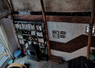 A cozy interior space with rustic and vintage design elements, featuring a black metal bookshelf filled with books and decorated with plants. The walls exhibit a weathered texture, and there are wooden beams adding to its rustic charm. A small blue stool and a fireplace with decorative items are placed nearby. The room has large windows with sheer curtains allowing natural light to enter. Various framed pictures or documents are displayed on the wall.