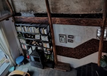 A cozy interior space with rustic and vintage design elements, featuring a black metal bookshelf filled with books and decorated with plants. The walls exhibit a weathered texture, and there are wooden beams adding to its rustic charm. A small blue stool and a fireplace with decorative items are placed nearby. The room has large windows with sheer curtains allowing natural light to enter. Various framed pictures or documents are displayed on the wall.