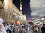 A large crowd of people dressed in traditional attire gathers outside a beautifully decorated mosque at dusk. The architecture is intricate with geometric patterns and large, open parasols providing shade. The sky is a gradient of deep blue to purple.