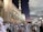 A large crowd of people dressed in traditional attire gathers outside a beautifully decorated mosque at dusk. The architecture is intricate with geometric patterns and large, open parasols providing shade. The sky is a gradient of deep blue to purple.