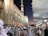 A large crowd of people dressed in traditional attire gathers outside a beautifully decorated mosque at dusk. The architecture is intricate with geometric patterns and large, open parasols providing shade. The sky is a gradient of deep blue to purple.