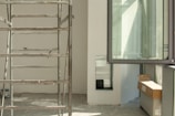 A construction or renovation site is visible, featuring metal scaffolding on a tiled floor. In the background, there's an open window allowing natural light to brighten the area. Cardboard boxes are placed on a surface, hinting at ongoing work. Electrical cables are scattered on the floor, and a cutout in the wall appears to be an unfinished part of the project.
