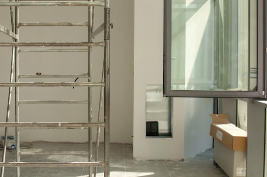 A construction or renovation site is visible, featuring metal scaffolding on a tiled floor. In the background, there's an open window allowing natural light to brighten the area. Cardboard boxes are placed on a surface, hinting at ongoing work. Electrical cables are scattered on the floor, and a cutout in the wall appears to be an unfinished part of the project.