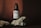 A dark-colored bottle with a pump dispenser sits among textured rocks. The label on the bottle reads 'Bro cosmetics' with descriptions of its uses as a double purpose balm, after shave, and stubble softener. The setting has a minimalist and earthy aesthetic with shadows creating a dramatic contrast.