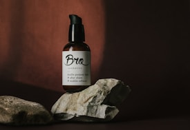 A dark-colored bottle with a pump dispenser sits among textured rocks. The label on the bottle reads 'Bro cosmetics' with descriptions of its uses as a double purpose balm, after shave, and stubble softener. The setting has a minimalist and earthy aesthetic with shadows creating a dramatic contrast.