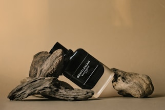 A bottle of men's fragrance labeled 'Gentleman Givenchy' is artfully positioned among pieces of driftwood, set against a warm beige background.