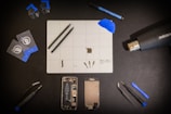 A meticulously organized workspace is shown with an assortment of tools and components arranged on a black surface. Central to the layout is a white grid mat that has various small parts and tools on it. Around the mat are a set of tweezers, suction cups, cards for prying, a blue spudger, a screwdriver, a heat gun, a marker, and two disassembled mobile phone parts, including a battery and screen. The general atmosphere is one of precision and technical activity.