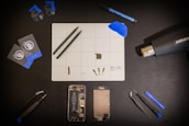 A meticulously organized workspace is shown with an assortment of tools and components arranged on a black surface. Central to the layout is a white grid mat that has various small parts and tools on it. Around the mat are a set of tweezers, suction cups, cards for prying, a blue spudger, a screwdriver, a heat gun, a marker, and two disassembled mobile phone parts, including a battery and screen. The general atmosphere is one of precision and technical activity.