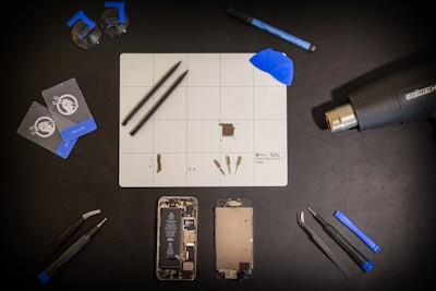 A meticulously organized workspace is shown with an assortment of tools and components arranged on a black surface. Central to the layout is a white grid mat that has various small parts and tools on it. Around the mat are a set of tweezers, suction cups, cards for prying, a blue spudger, a screwdriver, a heat gun, a marker, and two disassembled mobile phone parts, including a battery and screen. The general atmosphere is one of precision and technical activity.