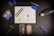 A meticulously organized workspace is shown with an assortment of tools and components arranged on a black surface. Central to the layout is a white grid mat that has various small parts and tools on it. Around the mat are a set of tweezers, suction cups, cards for prying, a blue spudger, a screwdriver, a heat gun, a marker, and two disassembled mobile phone parts, including a battery and screen. The general atmosphere is one of precision and technical activity.