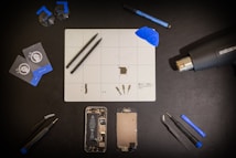 A meticulously organized workspace is shown with an assortment of tools and components arranged on a black surface. Central to the layout is a white grid mat that has various small parts and tools on it. Around the mat are a set of tweezers, suction cups, cards for prying, a blue spudger, a screwdriver, a heat gun, a marker, and two disassembled mobile phone parts, including a battery and screen. The general atmosphere is one of precision and technical activity.