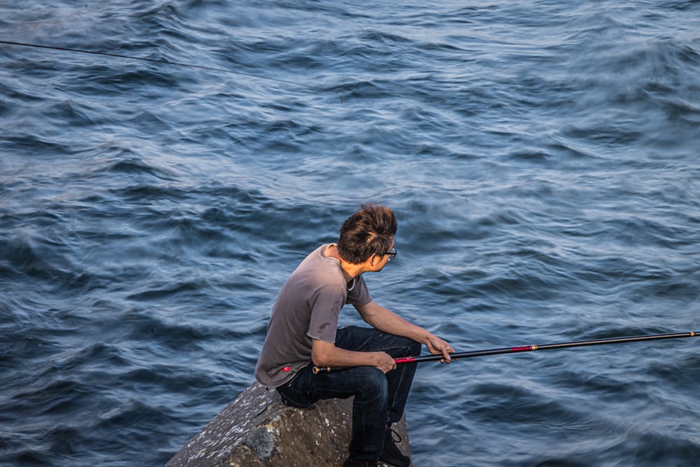 man in blue shirt and black pants sitting on rock in front of sea