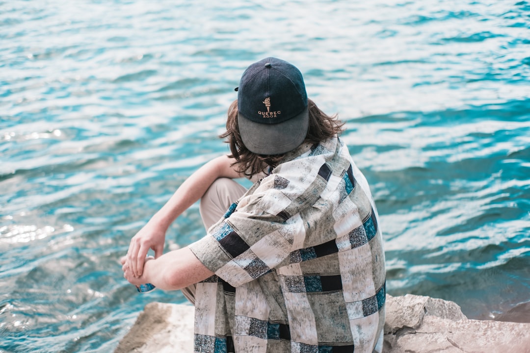 man in black cap and plaid shirt sitting on rock near body of water during daytime