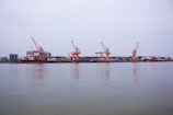 A dockyard with multiple large cranes positioned along a waterfront, handling stacked shipping containers of various colors. The calm water reflects part of the scene, with overcast skies creating a serene atmosphere.