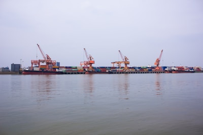 A dockyard with multiple large cranes positioned along a waterfront, handling stacked shipping containers of various colors. The calm water reflects part of the scene, with overcast skies creating a serene atmosphere.