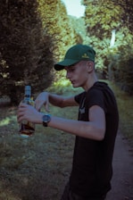 A young person in a black shirt and green cap stands outdoors, holding a bottle of amber liquid. They appear to be in a forested area with sunlit trees and a dirt path. Their expression is focused, and they're wearing a watch on their wrist.