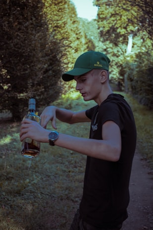 A young person in a black shirt and green cap stands outdoors, holding a bottle of amber liquid. They appear to be in a forested area with sunlit trees and a dirt path. Their expression is focused, and they're wearing a watch on their wrist.