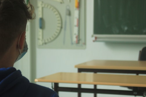 A person wearing a mask sits in a classroom, facing forward. In the background, there are empty desks, a green chalkboard, and classroom tools such as a protractor and meter ruler hanging on the wall.