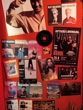 A vibrant collage of music posters and promotional materials is displayed on a red wall. The collection features various artists and bands, with images and text highlighting events and albums. In the mix, there are colorful and black-and-white posters, including an image of someone holding a trophy and a vinyl record affixed to the wall. The aesthetic is lively and energetic, with a focus on musical culture.