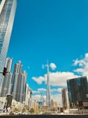 A vibrant cityscape features a collection of modern skyscrapers, with a tall, iconic tower at the center. The buildings are under a bright blue sky accented by scattered white clouds. The foreground includes a street with traffic lights and a few visible cars and pedestrians.