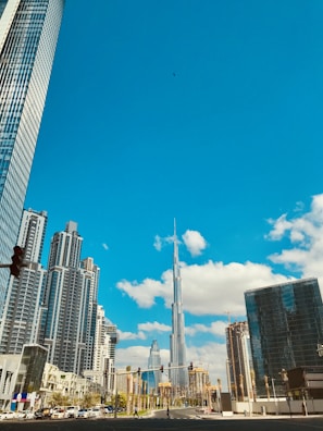 A vibrant cityscape features a collection of modern skyscrapers, with a tall, iconic tower at the center. The buildings are under a bright blue sky accented by scattered white clouds. The foreground includes a street with traffic lights and a few visible cars and pedestrians.