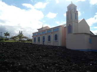 A light-colored church with pastel accents stands beside a dark rocky landscape. The structure features arched windows and a bell tower, set against a backdrop of a bright blue sky with scattered clouds. Trees and greenery are visible in the background.