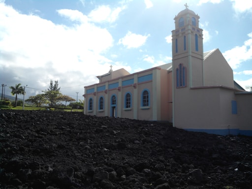 A light-colored church with pastel accents stands beside a dark rocky landscape. The structure features arched windows and a bell tower, set against a backdrop of a bright blue sky with scattered clouds. Trees and greenery are visible in the background.
