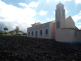 A light-colored church with pastel accents stands beside a dark rocky landscape. The structure features arched windows and a bell tower, set against a backdrop of a bright blue sky with scattered clouds. Trees and greenery are visible in the background.