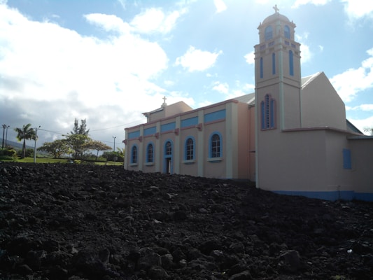 A light-colored church with pastel accents stands beside a dark rocky landscape. The structure features arched windows and a bell tower, set against a backdrop of a bright blue sky with scattered clouds. Trees and greenery are visible in the background.