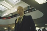 A mannequin dressed in a formal suit and tie is positioned inside a spacious, modern building with high ceilings. The lighting is soft and ambient, with fluorescent lights above. Shelving or balcony spaces are visible, suggesting an indoor shopping or exhibition area.