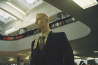 A mannequin dressed in a formal suit and tie is positioned inside a spacious, modern building with high ceilings. The lighting is soft and ambient, with fluorescent lights above. Shelving or balcony spaces are visible, suggesting an indoor shopping or exhibition area.