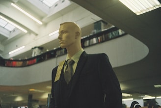 A mannequin dressed in a formal suit and tie is positioned inside a spacious, modern building with high ceilings. The lighting is soft and ambient, with fluorescent lights above. Shelving or balcony spaces are visible, suggesting an indoor shopping or exhibition area.