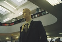 A mannequin dressed in a formal suit and tie is positioned inside a spacious, modern building with high ceilings. The lighting is soft and ambient, with fluorescent lights above. Shelving or balcony spaces are visible, suggesting an indoor shopping or exhibition area.